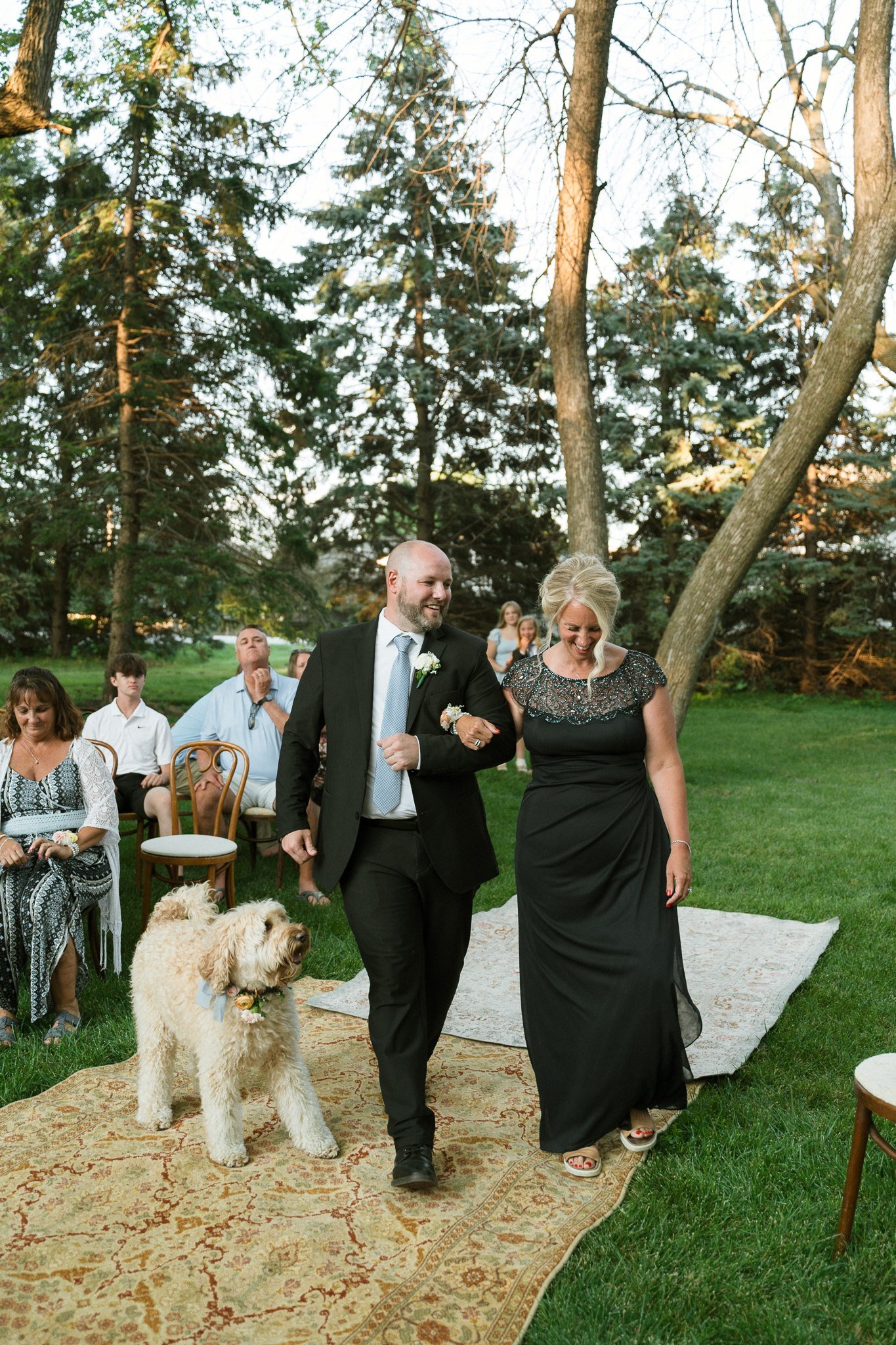 Dog walking the outdoor aisle with wedding guests in a black suit and beaded dress