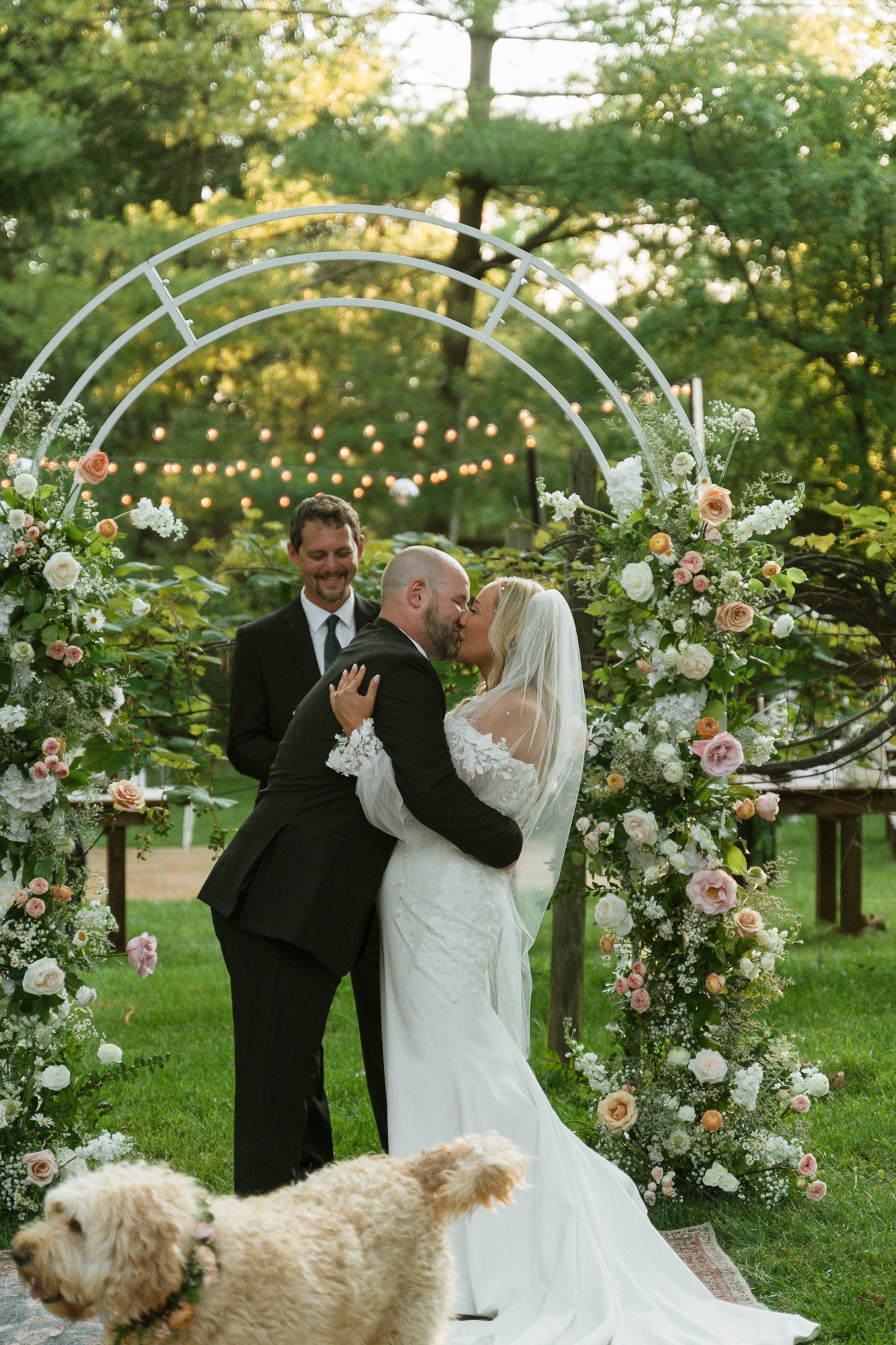Bride and groom sharing their first kiss under a floral arch with their dog in the foreground