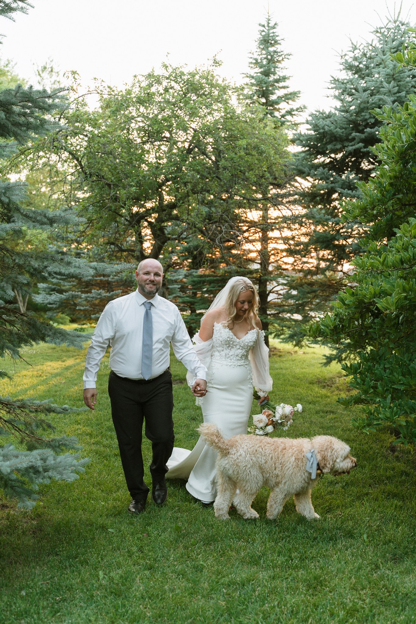 Bride and groom walking with their dog wearing a blue bow on golden-hour portraits
