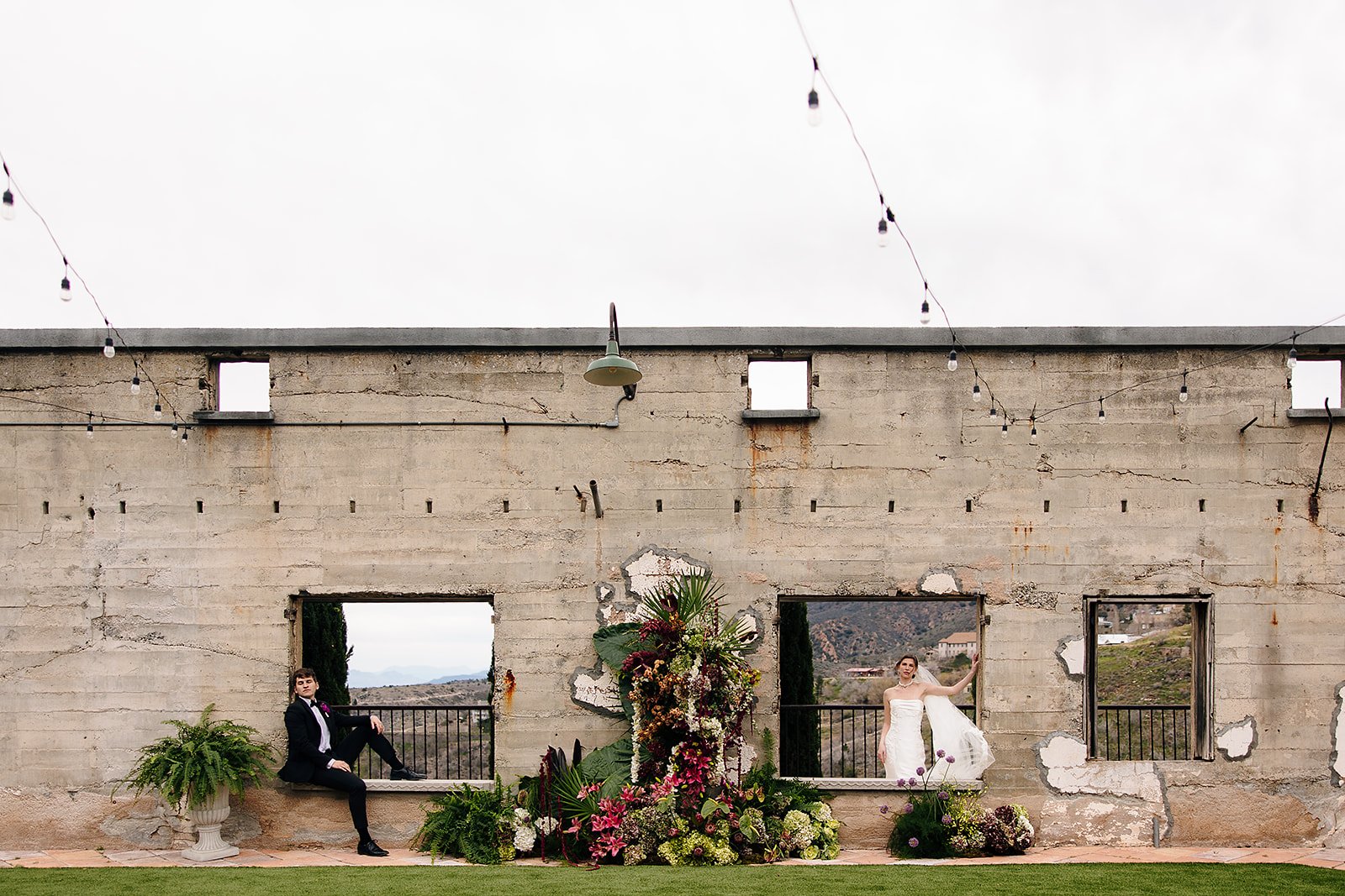 Couple photographed in the window openings of a ruined concrete wall with a floral installation